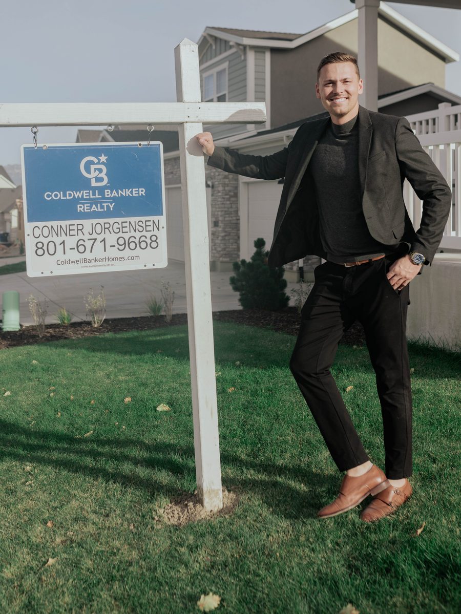 Conner leaning on his Coldwell Banker for-sale sign in front of a listed home